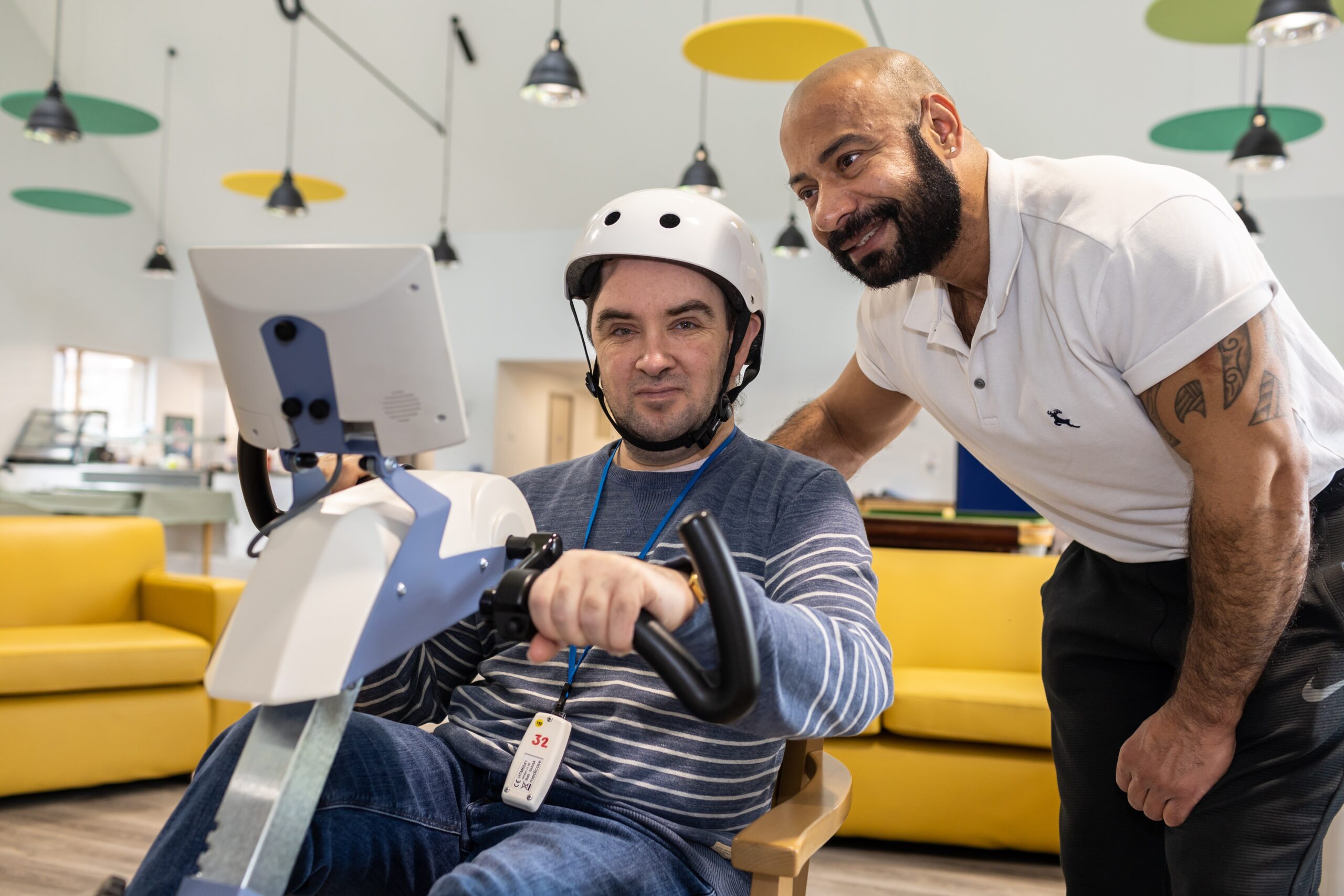 man on an exercise bike wearing a white helmet, supported by a therapist