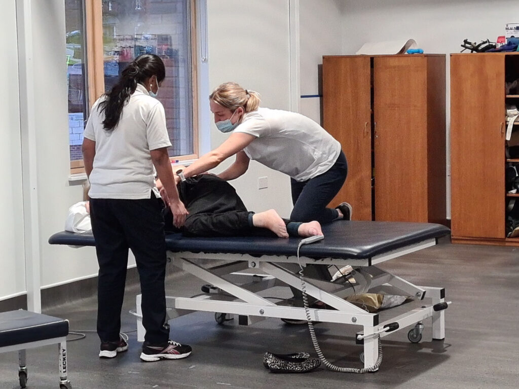2 physiotherapists support a client to do a leg exercise whilst lying on a therapy bed
