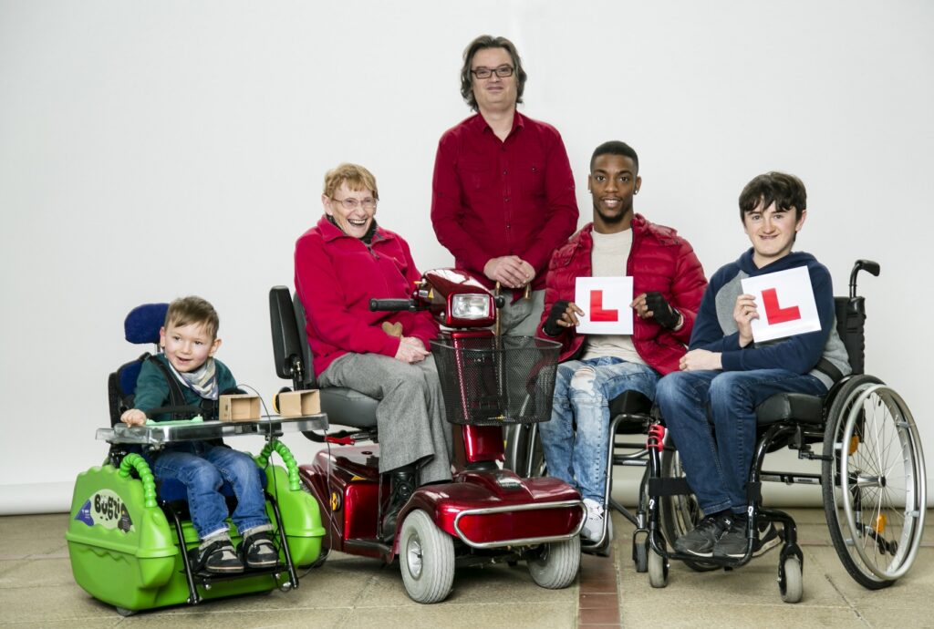 5 people of all ages with wheelchairs, scooters or mobility aids. Two young adults holding L plates