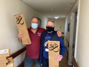 Two people, wearing face masks, standing in a corridor. They are both holding cardboard flower boxes.