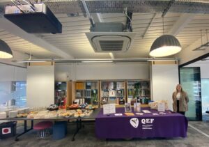 A QEF fundraising stall set out in a room. The table is dressed in a purple tablecloth, and there are publicity materials on the table.