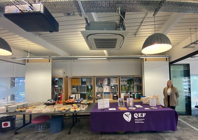 A QEF fundraising stall set out in a room. The table is dressed in a purple tablecloth, and there are publicity materials on the table.