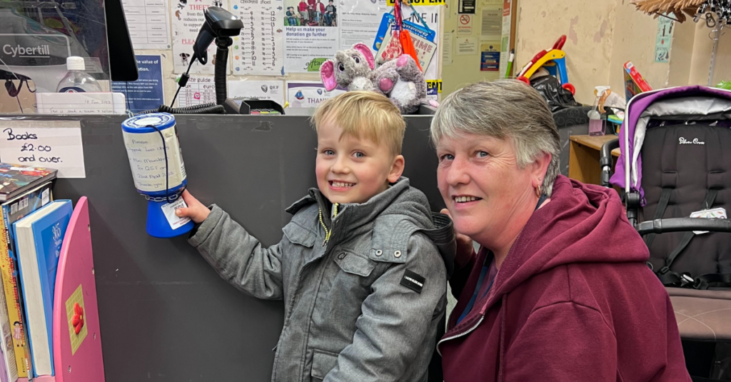 Leo is a four year old boy, he is blonde and is grinning big whilst holding a blue charity collection pot. Next to him is a lady with grey hair, she is smiling too