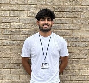 Kishan is wearing a white t-shirt with an ID badge on in front of a wall