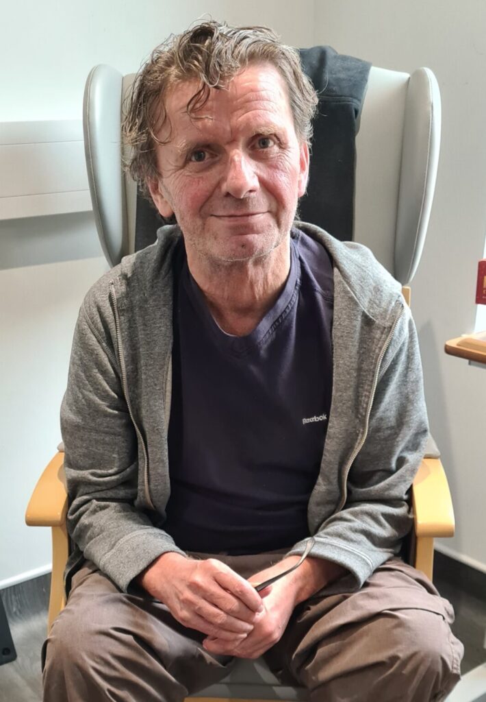 Gareth sitting in an armchair chair looking to camera, wearing a blue t-shirt and grey hoodie