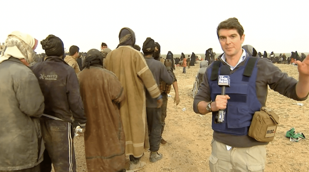 Benjamin standing in a desert location, looking to camera wearing a safety vest, with multiple other people walking away from him on the left, wearing head coverings and long brown coats