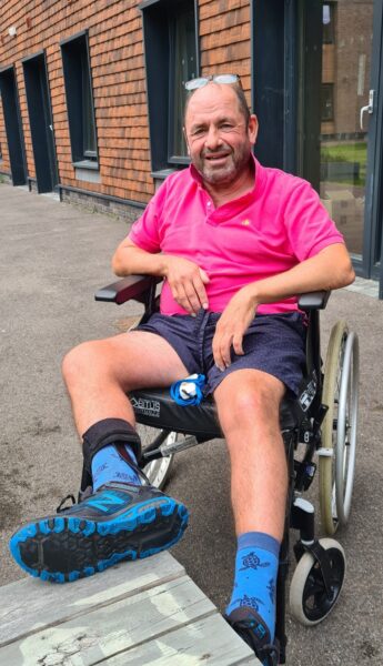 Andrew has his glasses on his head and he is sitting in a wheelchair outside the CRC, with one leg balanced on a bench in front of him.