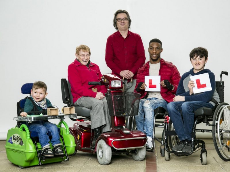 5 people of all ages with wheelchairs, scooters or mobility aids. Two young adults holding L plates