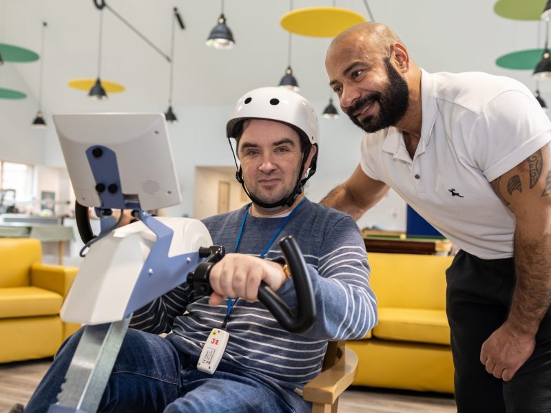 Male client wearing a white helmet sitting on an exercise bike, supported by a member of staff