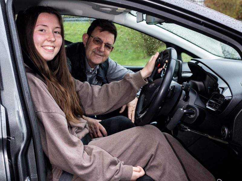 A young lady sitting in the driving seat of a car, smiling, with the door open, and a driving instructor smiling in the passenger seat