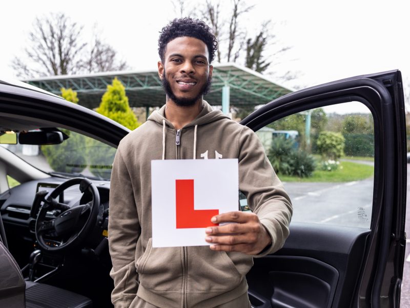 Young man smiling and standing beside an open car door with an L plate in his hand