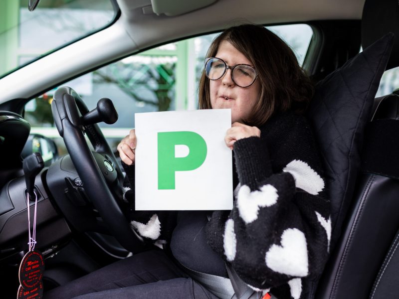 lady in a driving seat of a car holding a green P plate