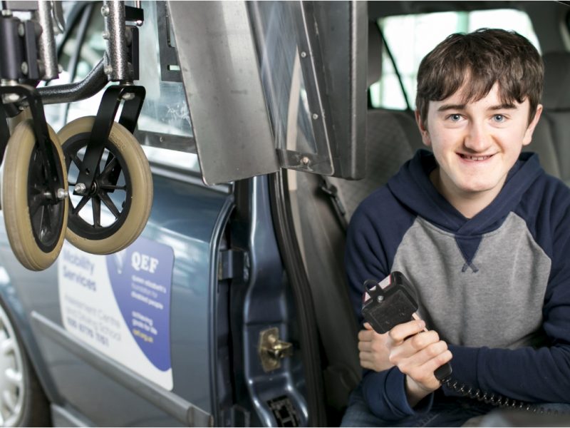 young man sitting in a car seat, holding a remote which he is using to lower a wheelchair from the roof of the car