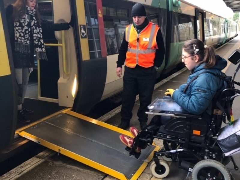 A person getting onto a train, using a ramp to steer their wheelchair up. A train guard in a high viz jacket stands next to the ramp, and a third person is stood in the doorway.